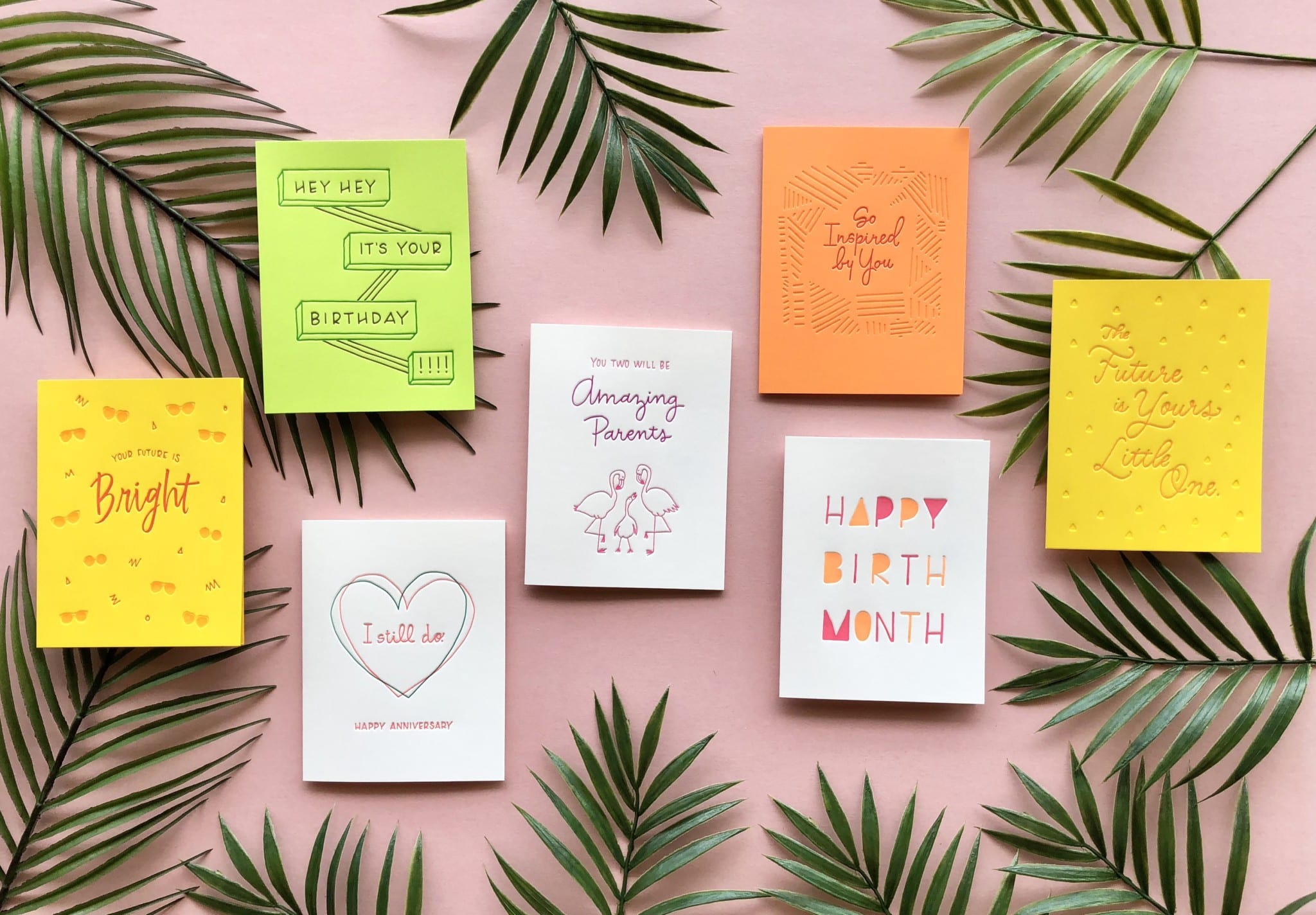 assorted greeting cards arranged on light pink background with palm leaves
