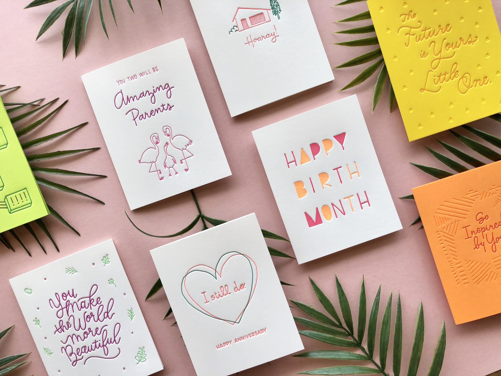 Colorful greeting cards arranged on a light pink background with palm leaves.