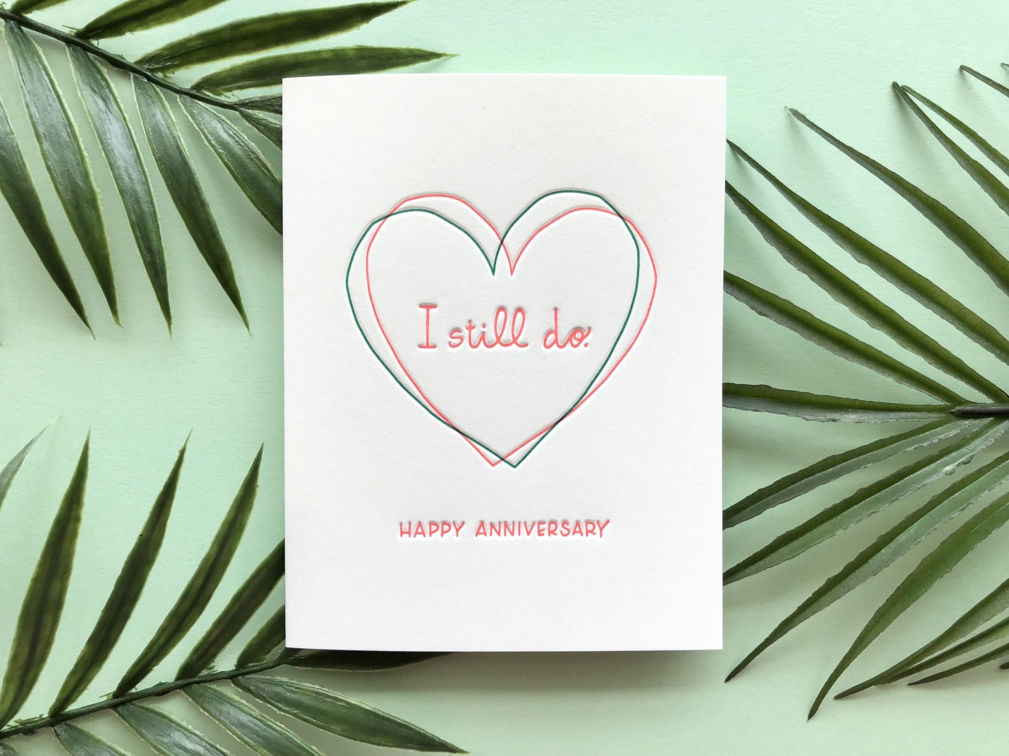 Greeting card with a hand-drawn heart and the message "I still do - Happy Anniversary" on a green background with palm leaves.