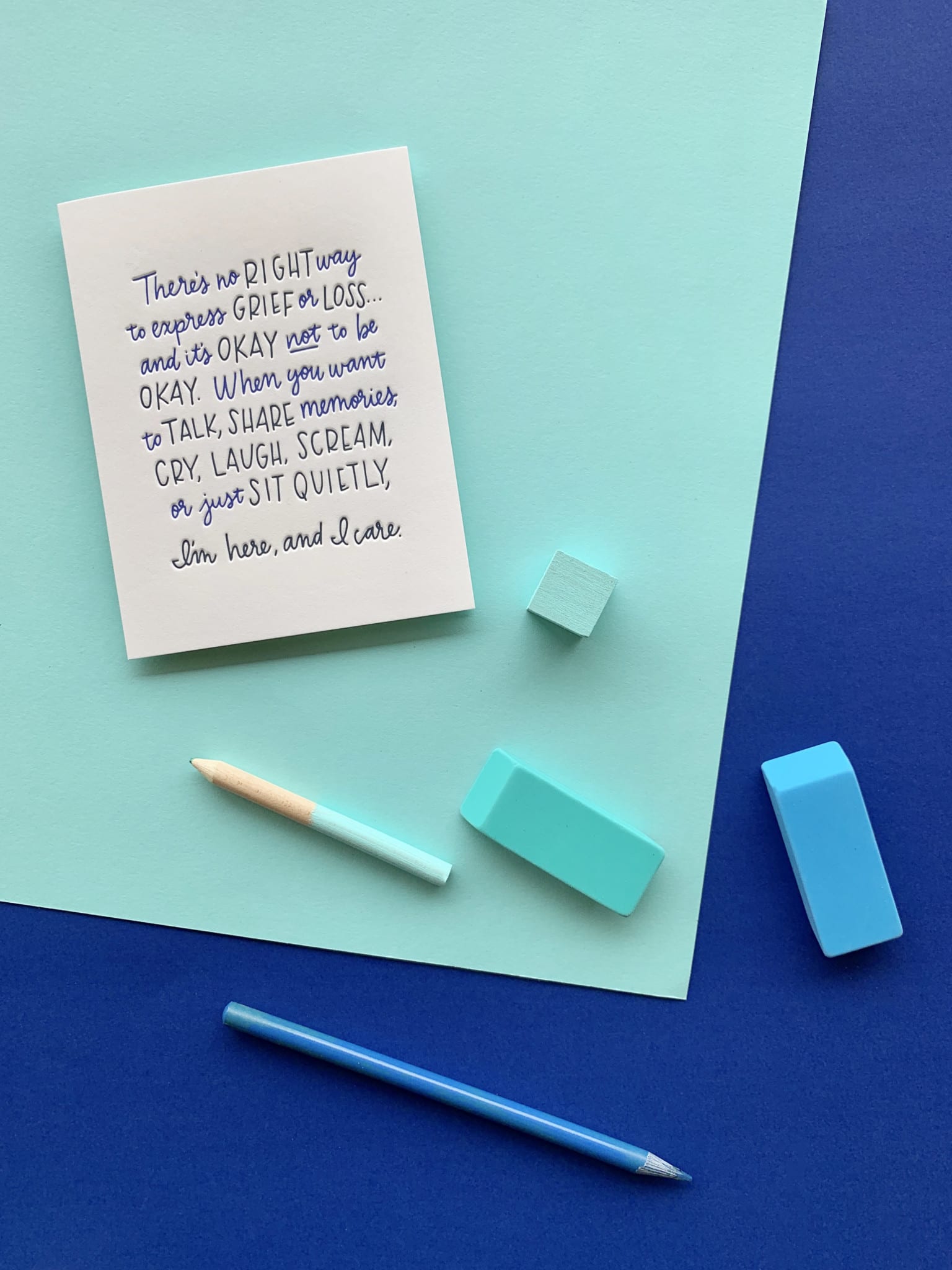 Words of encouragement for times of grief cover this greeting card. The composition in completed by several blue color desk objects spread over a multi-layered paper background.