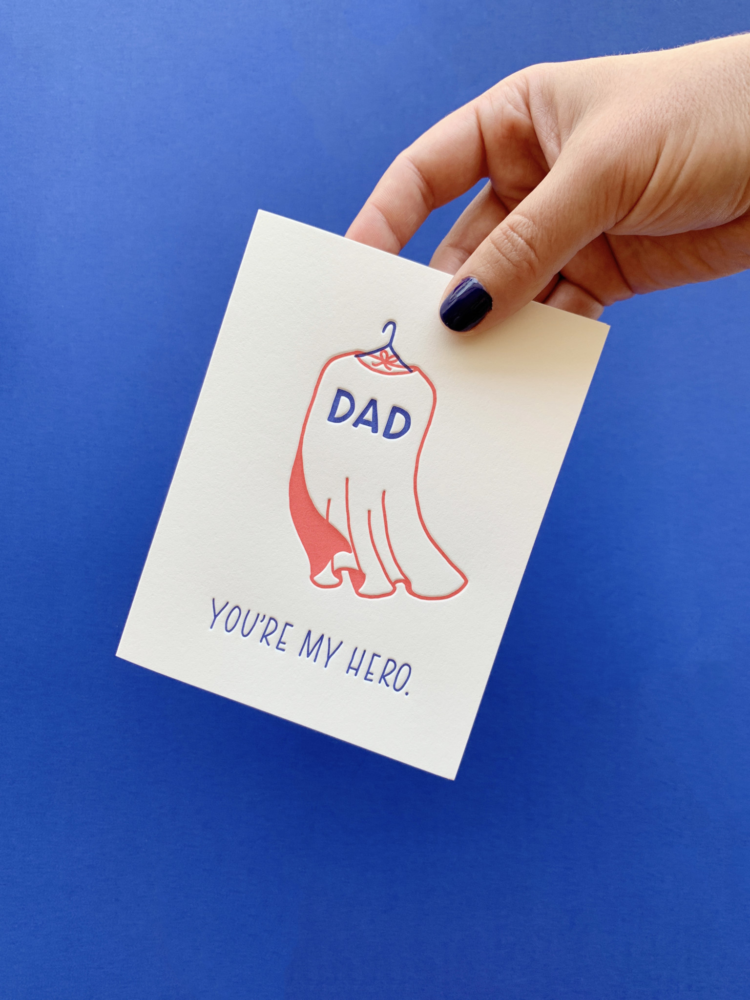 Dad, You're My Hero card held by hand with blue painted nails on a blue background.