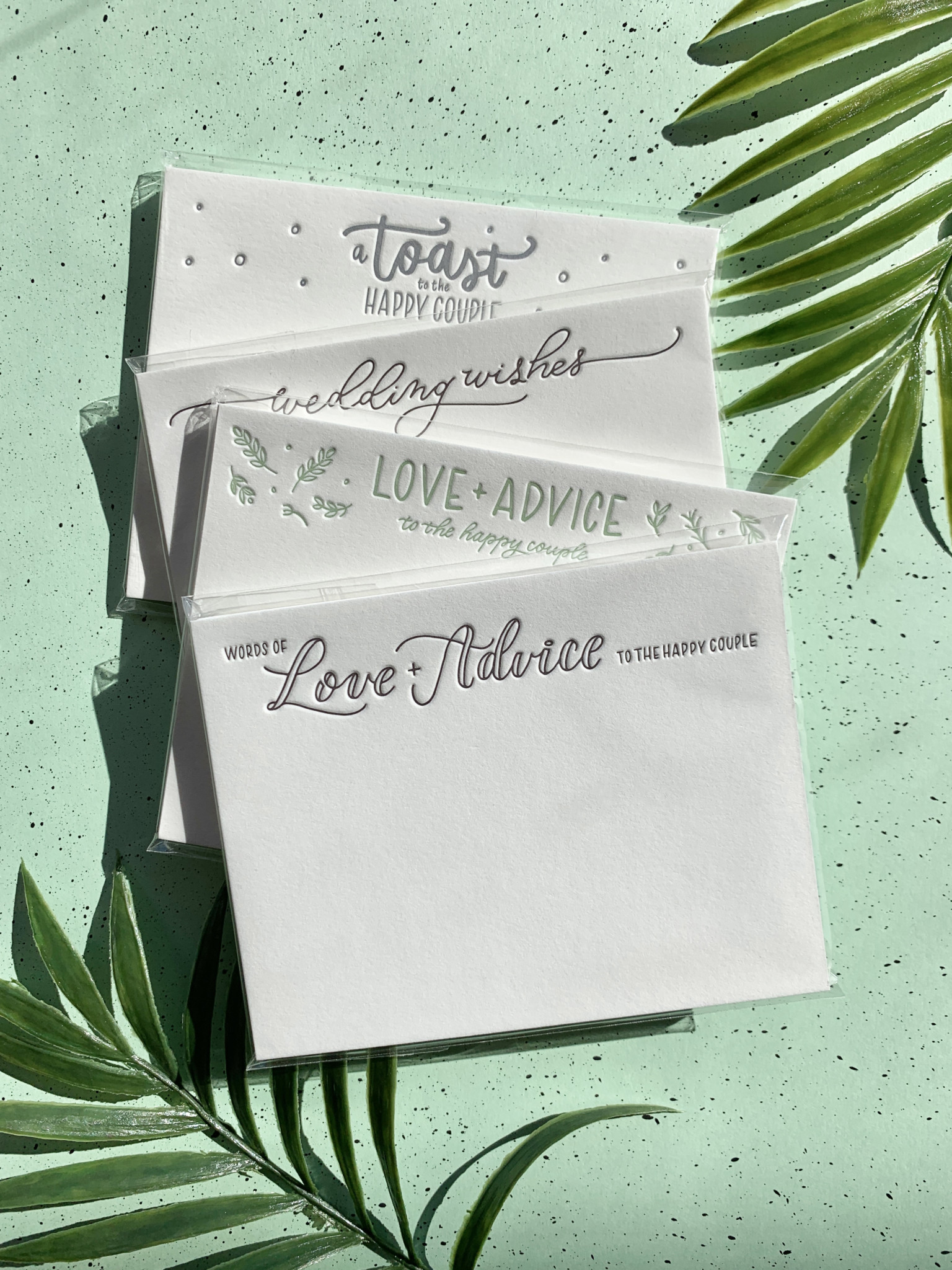 Soft packs of letterpress printed wedding advice cards arranged on a green paper background with palm leaf accents.