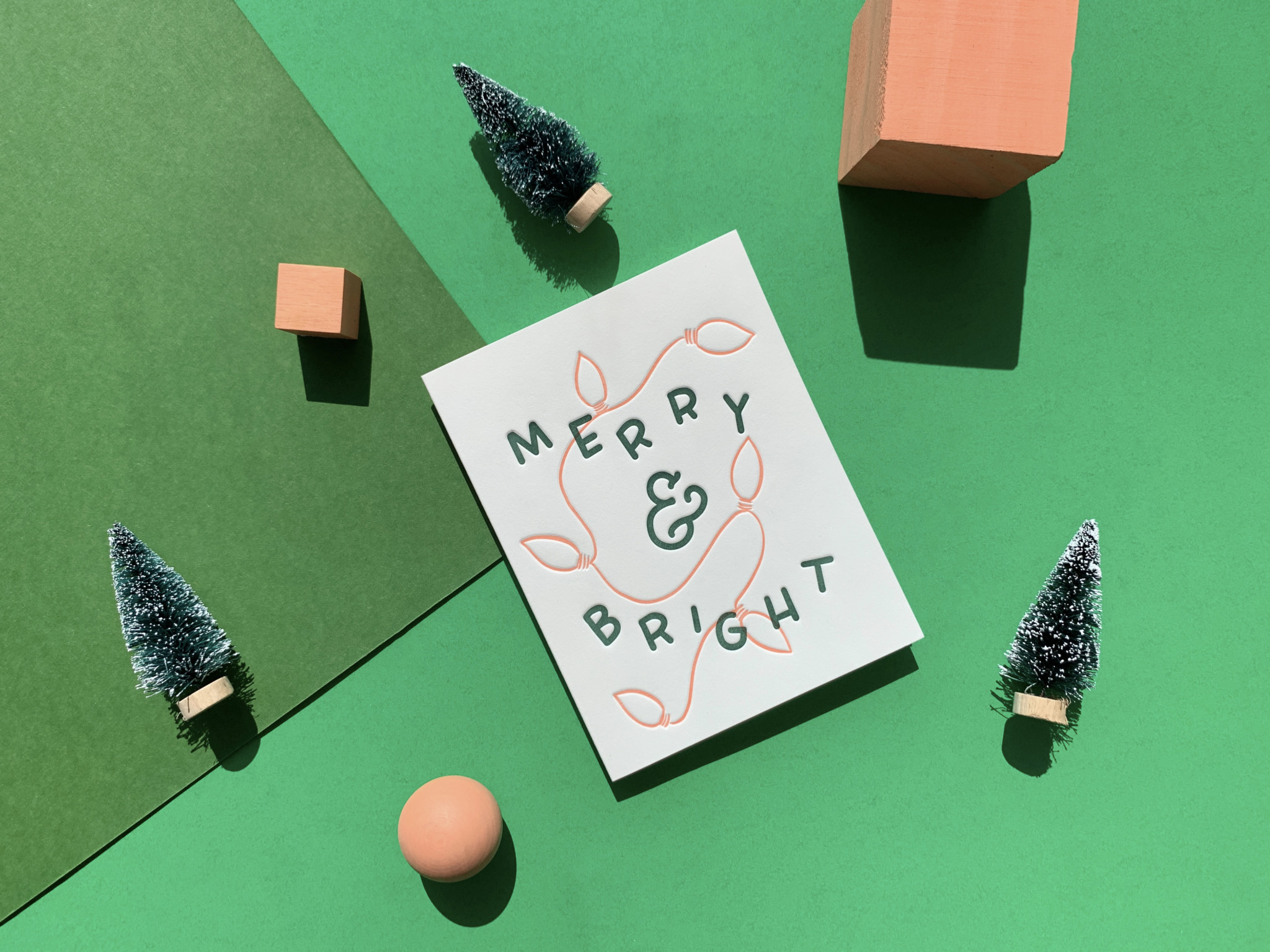Greeting card reads "Merry & Bright" with illustrations of lights. Styled with holiday objects on a layered paper background.