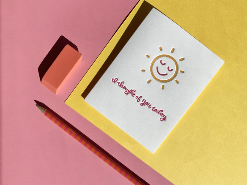 Greeting card featuring smiling sunshine is positioned against pink and yellow paper background with pink pencil and eraser also visible