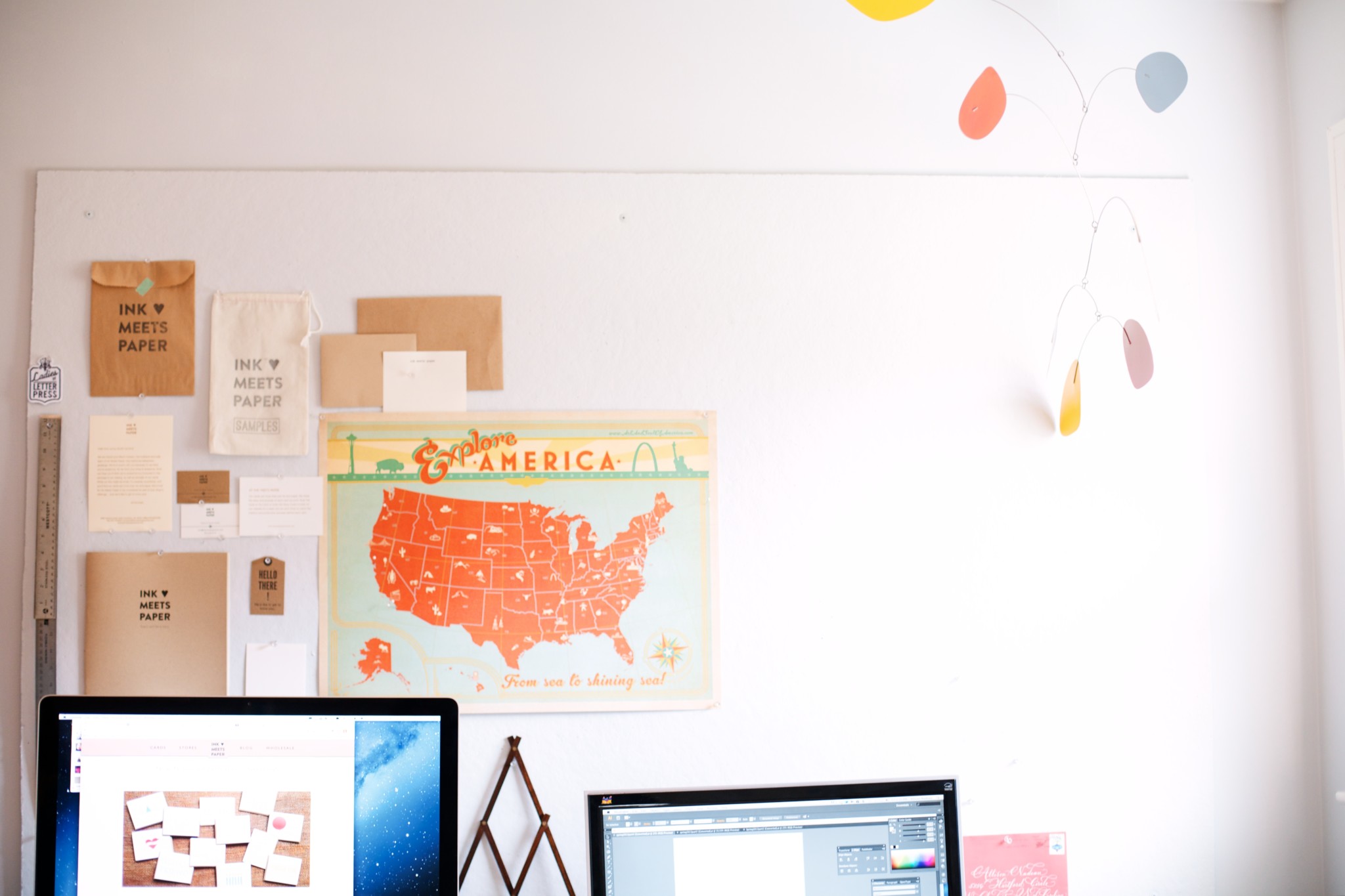 Two computer monitors are in the foreground with INK MEETS PAPER brand collateral pinned to the wall in the background along with a map of the United States
