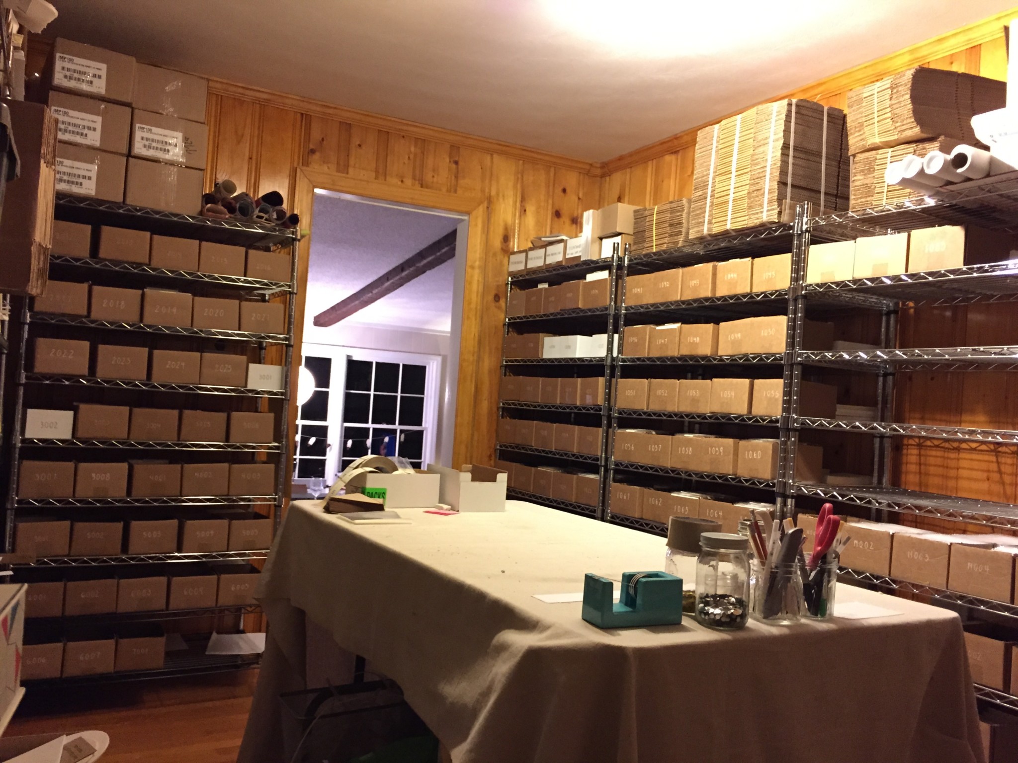Floor-to-ceiling metal shelves are filled with brown boxes containing greeting card inventory, and a table for assembly sits in the middle of the small room.
