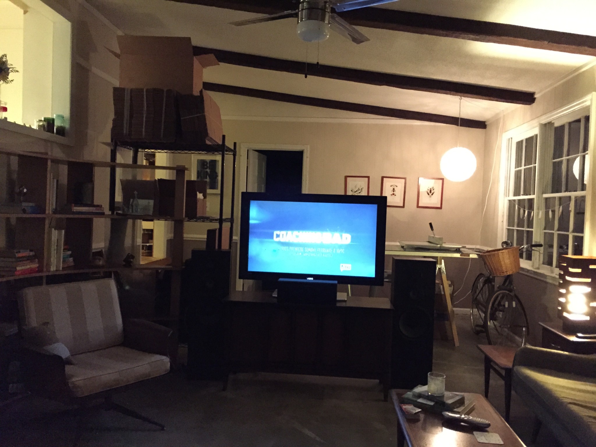 TV, chair, bookcase, and couch are positioned in one-half of the living room with a work table and inventory shelf against the remainder of the room