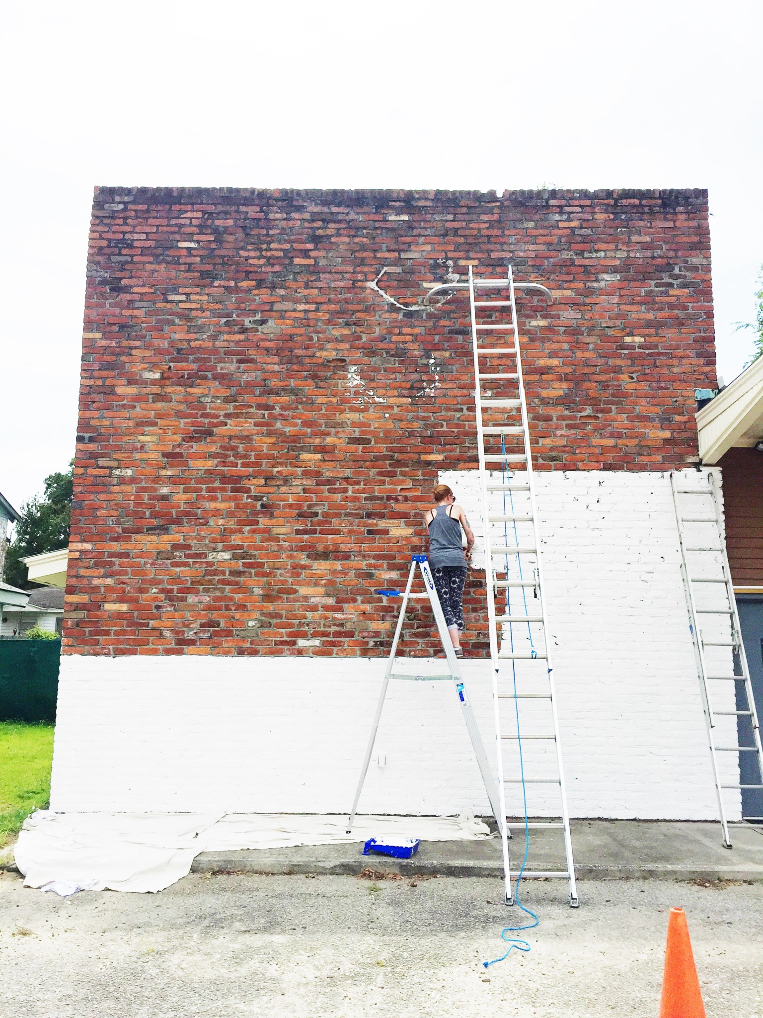 Brick wall of studio is being painted bright white by a female and two large ladders are propped against wall