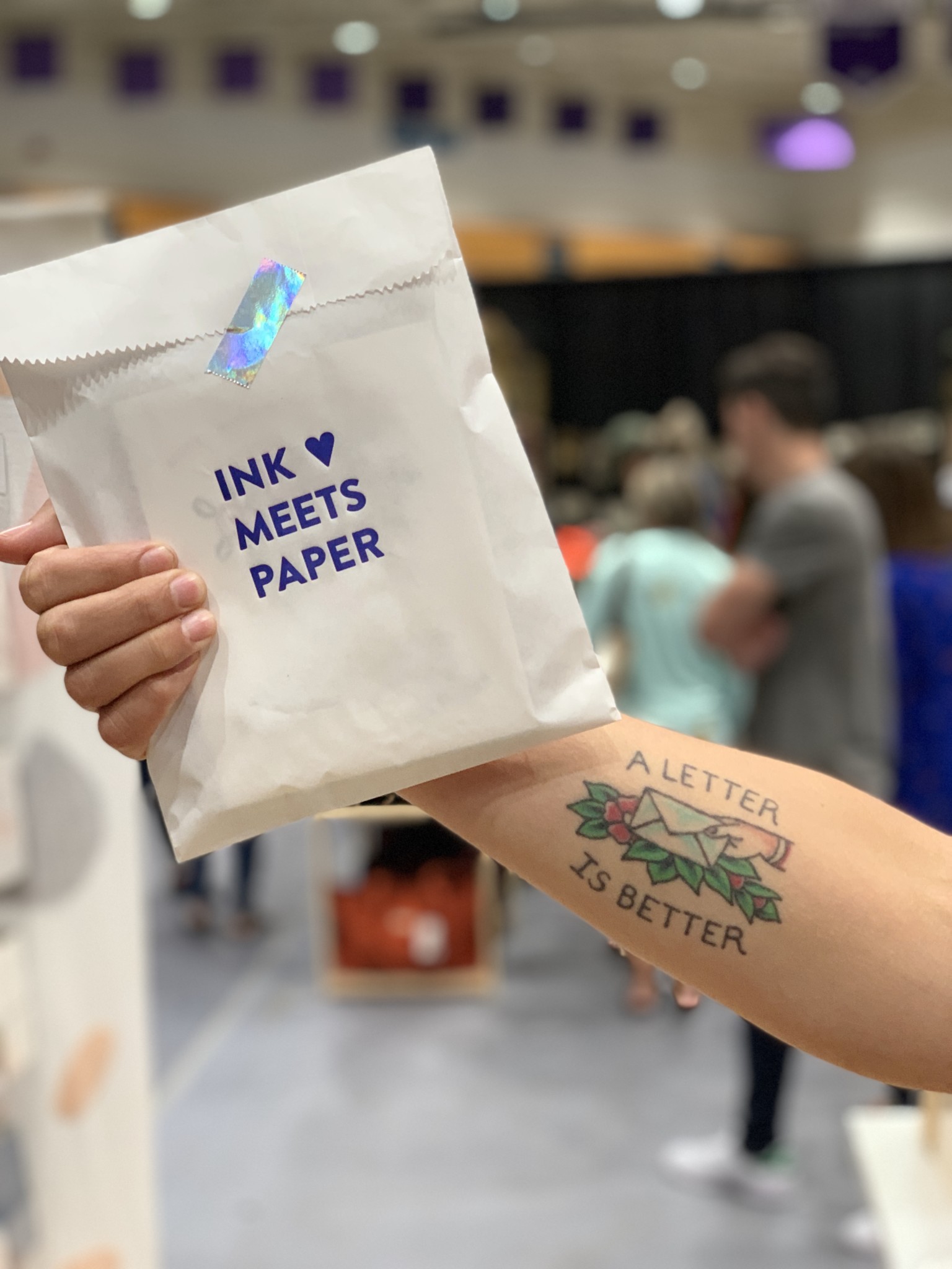 INK MEETS PAPER merchandise bag is held by a hand that shows the arm with a tattoo that depicting a letter held in a hand with the text "A Letter is Better"