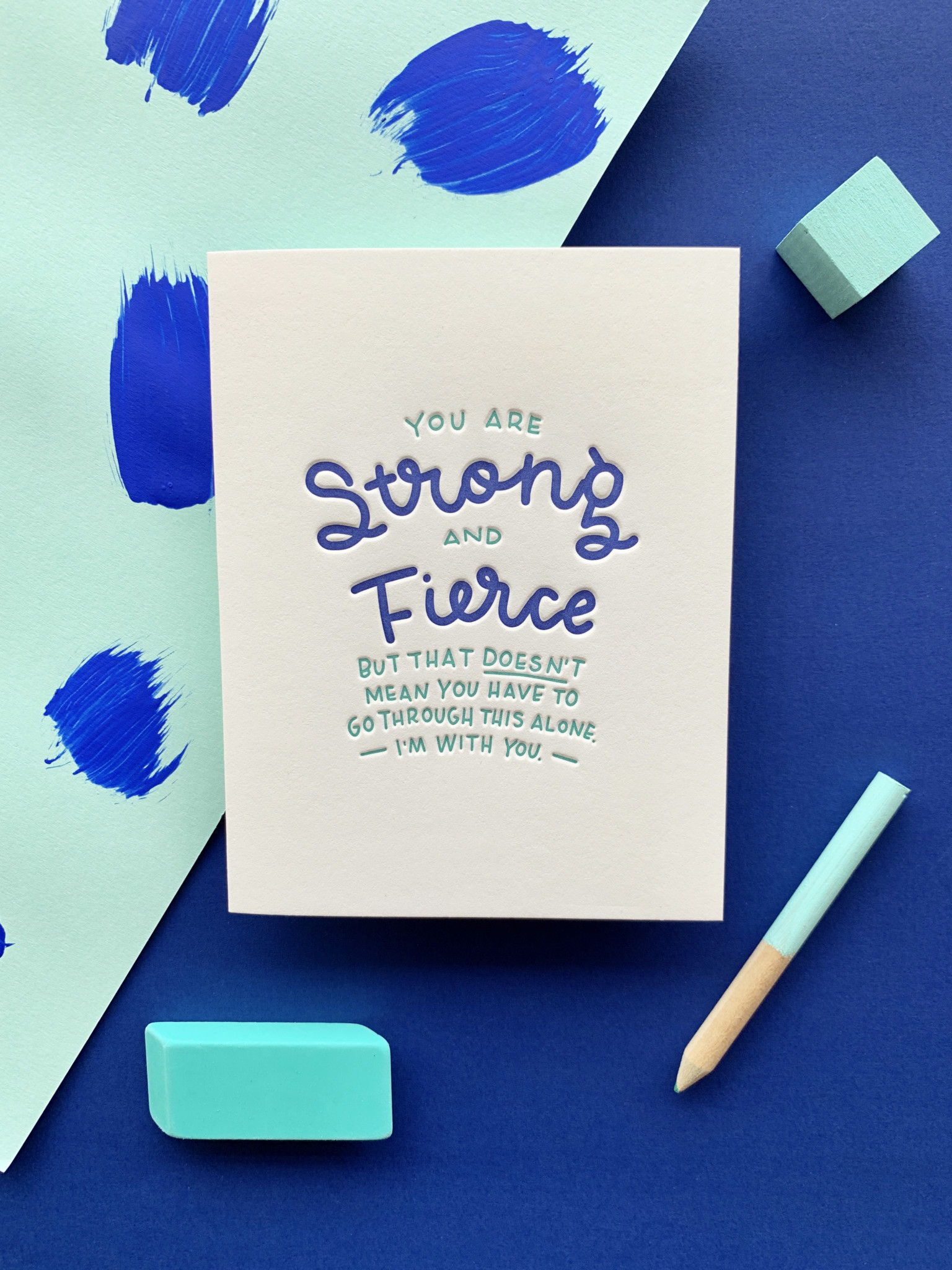 Greeting card features hand-lettered sentiment that reads "You are strong and fierce by that doesn't mean you have to go through this along. I'm with you." Card is positioned on a teal and bright blue paper background.