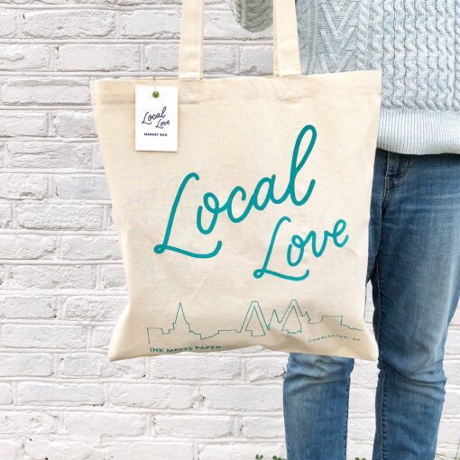 Model holding market bag. Bag shows handlettered words Local Love and a line drawing of the skyline of Charleston, SC