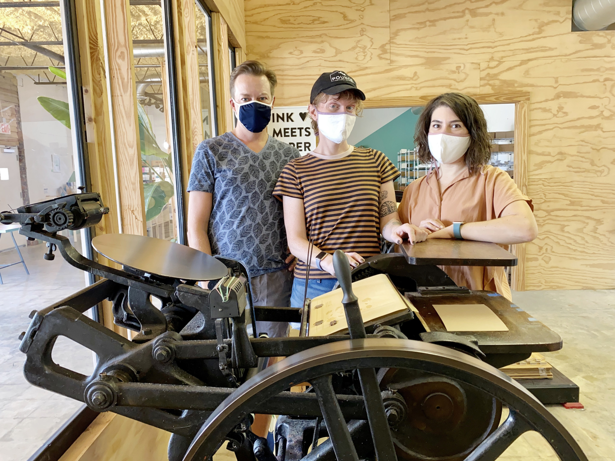 Members of the INK MEETS PAPER Team posing behind a letterpress printing machine. Each person is wearing a mask per COVID-19 CDC Guidelines.