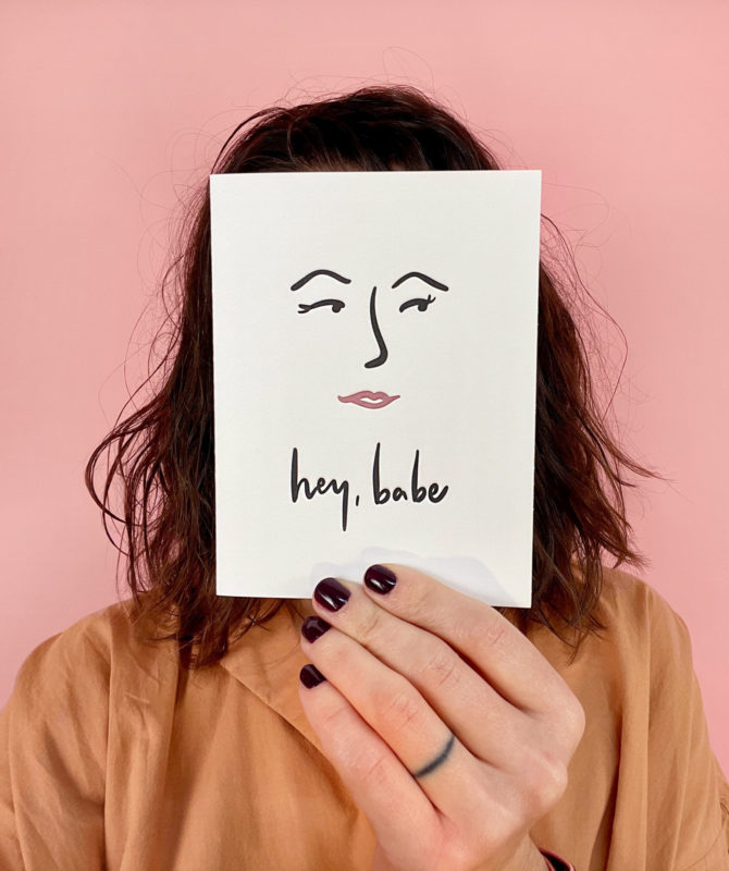 woman holding card with drawn face that reads "hey, babe" in front of her face